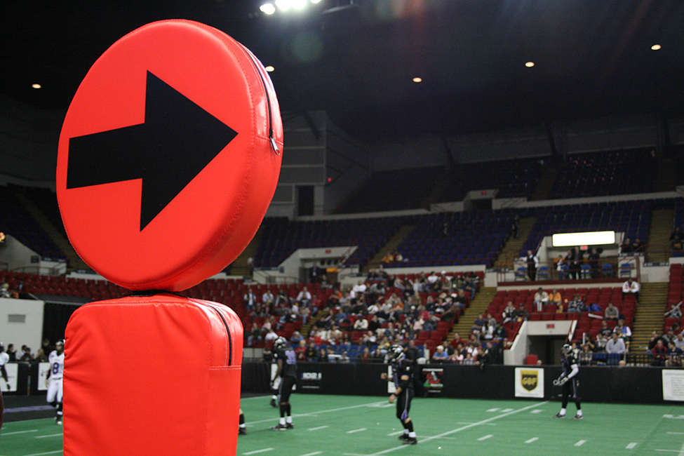 A large orange circle with an arrow printed on it facing to the right atop a rectangular base, known as chains in football, is shown prominently in the foreground.  In the background there is a game of football ongoing with fans in their seats.