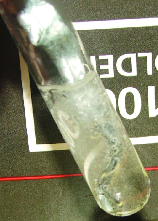 A glass tube holding a metallic solid in a colorless liquid is shown laying on a black background with white lettering.