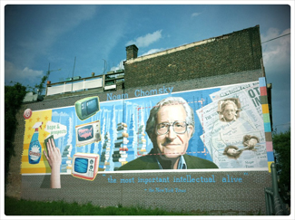 A photograph shows a mural on the side of a building. The mural includes Chomsky's face, along with some newspapers, televisions, and cleaning products. At the top of the mural, it reads “Noam Chomsky.” At the bottom of the mural, it reads “the most important intellectual alive.”