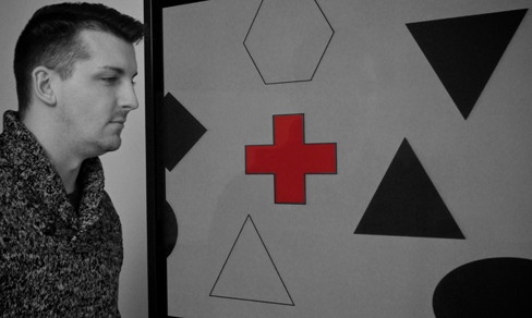 A photograph shows a person staring at a screen that displays one red cross toward the left side and numerous black and white shapes all over.